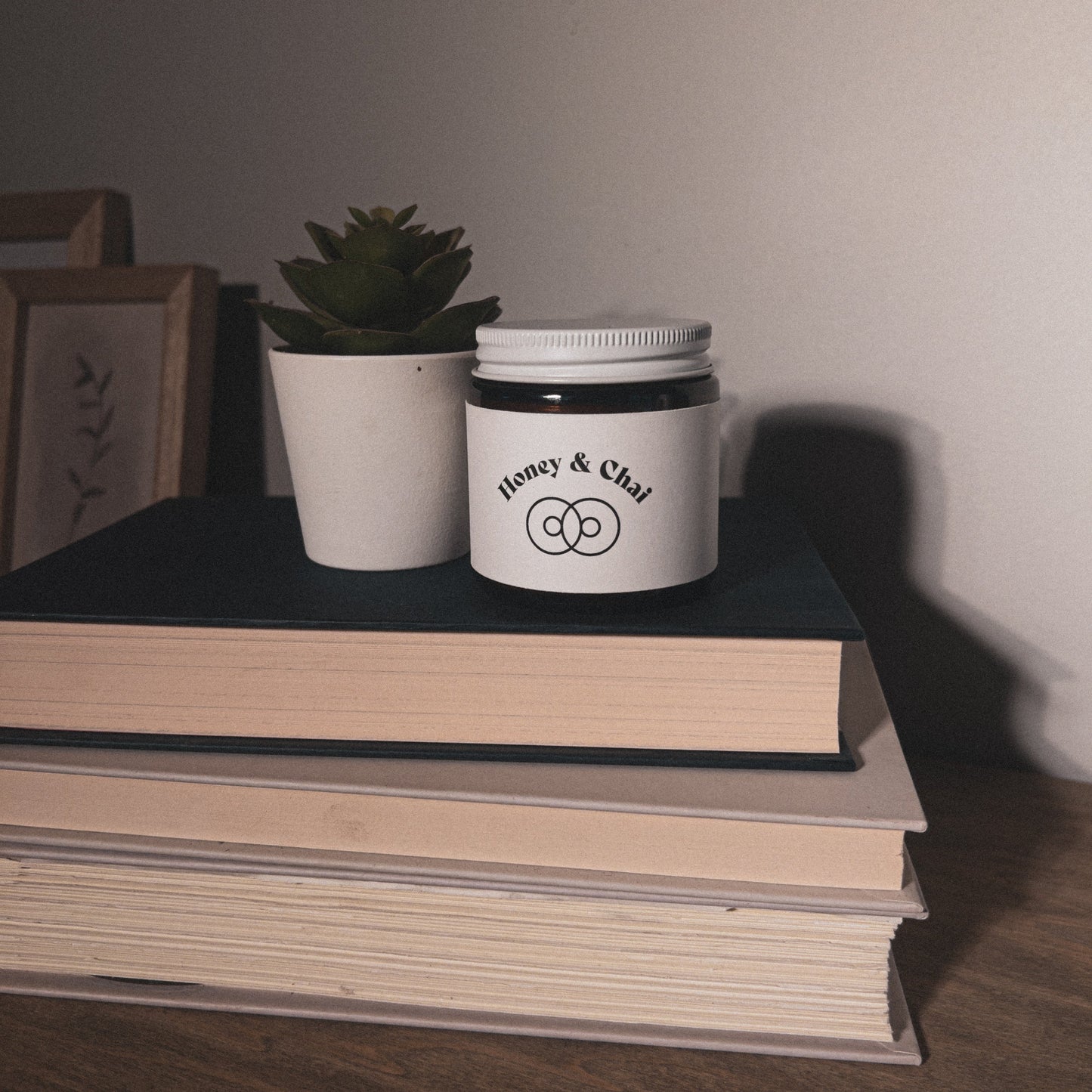 Honey & Chai Scented Beeswax Candle 4 oz in amber glass jar with white lid. Candle on stack of books with potted succulent and framed pictures.